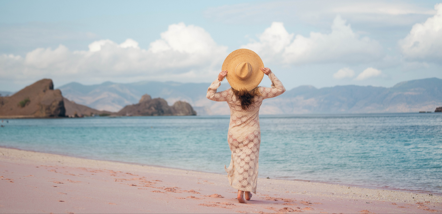 Annie in a dress and hat standing on a beach with mountains in the background